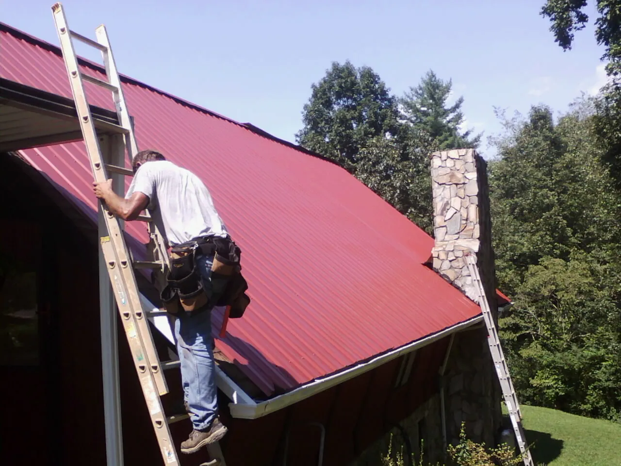 Roofer on a ladder beside a red metal roof and stone chimney — Soffit & Fascia Repair by Roofing Brothers in Washington Navy Yard